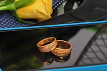 Two gold rings engraved with names are placed on a reflective surface, likely a smartphone screen. In the background, a yellow rose with green leaves and a textured blue fabric can be seen.
