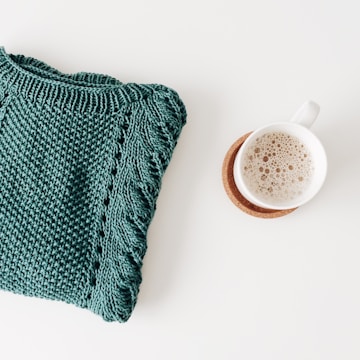 A folded green knitted sweater lies beside a cup of coffee with frothy milk, both placed on a light surface. The coffee cup sits on a cork coaster, adding a rustic element to the cozy setting.