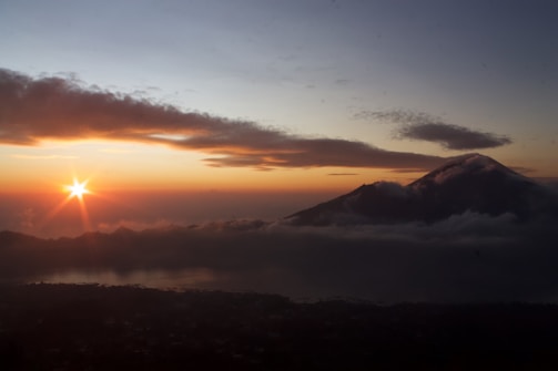 A serene landscape with a mountain partially shrouded in clouds, silhouetted against a vibrant sunrise. The sky is a gradient of soft blues transitioning into deep oranges and yellows as the sun peeks over the horizon. Gentle cloud formations are spread across the sky, adding depth to the scene.