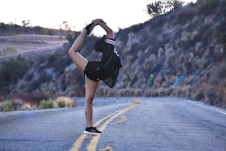 A person performs an advanced yoga or dance pose, balancing on one leg while holding the other leg in the air. They are on a deserted, curved road surrounded by hills covered in dry grass and sparse foliage. The scene is set during what appears to be early morning or late afternoon, with soft lighting.