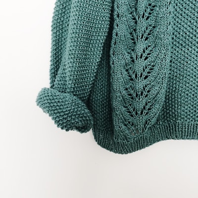 A knitted teal sweater with a detailed pattern, featuring a rolled-up sleeve. The texture is prominent, showcasing the intricate design and soft material.