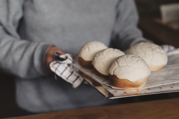 A person holding a metal tray with four freshly baked buns topped with a crackly crust. The person is wearing a grey sweater and is using a checkered cloth to handle the tray.
