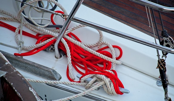 Bright red and off-white ropes are coiled and entangled on the deck of a boat. The area is set against polished metal fittings and wooden decking, suggesting a nautical setting.