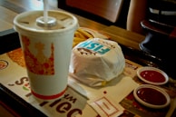 A fast food meal on a tray consisting of a wrapped sandwich, a disposable cup with a straw, some french fries, and two small containers of ketchup. There are also some napkins placed on the tray. Part of a person's arm is visible in the background, along with a handbag on the table.