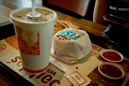 A fast food meal on a tray consisting of a wrapped sandwich, a disposable cup with a straw, some french fries, and two small containers of ketchup. There are also some napkins placed on the tray. Part of a person's arm is visible in the background, along with a handbag on the table.