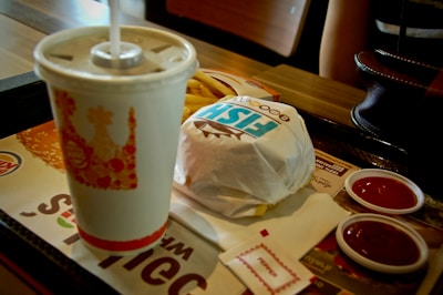 A fast food meal on a tray consisting of a wrapped sandwich, a disposable cup with a straw, some french fries, and two small containers of ketchup. There are also some napkins placed on the tray. Part of a person's arm is visible in the background, along with a handbag on the table.