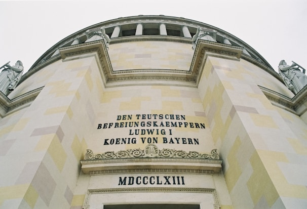 A grand architectural structure with a detailed facade, featuring classical columns and statues on top. The walls display inscribed text in a bold, elegant font. The large letters spell out phrases in German, highlighting the historical importance.