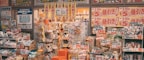 A cluttered storefront of a Japanese retail shop filled with various products, including snacks, groceries, and household items. Brightly colored sale signs in Japanese advertise discounts and promotions, creating a busy and vibrant atmosphere. The shelves and baskets are packed, and the overall space has a crowded appearance.