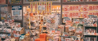 A cluttered storefront of a Japanese retail shop filled with various products, including snacks, groceries, and household items. Brightly colored sale signs in Japanese advertise discounts and promotions, creating a busy and vibrant atmosphere. The shelves and baskets are packed, and the overall space has a crowded appearance.