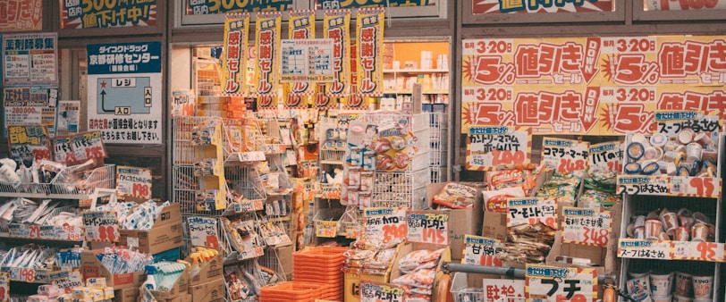 A cluttered storefront of a Japanese retail shop filled with various products, including snacks, groceries, and household items. Brightly colored sale signs in Japanese advertise discounts and promotions, creating a busy and vibrant atmosphere. The shelves and baskets are packed, and the overall space has a crowded appearance.
