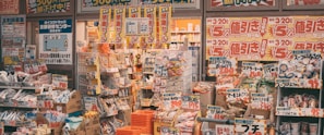 A cluttered storefront of a Japanese retail shop filled with various products, including snacks, groceries, and household items. Brightly colored sale signs in Japanese advertise discounts and promotions, creating a busy and vibrant atmosphere. The shelves and baskets are packed, and the overall space has a crowded appearance.