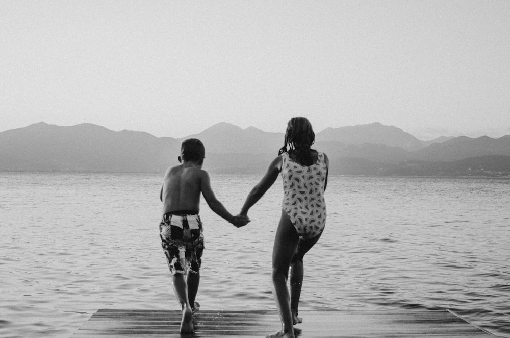 grayscale photography of girl and boy standing beside body of water