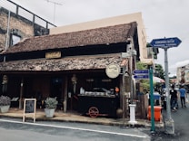 A rustic, traditional building with a sloping tiled roof and wooden exterior sits at a street corner. A signboard with foreign script hangs above the entrance. Several potted plants and a chalkboard menu frame the doorway, suggesting a cafe or small restaurant. Street signs in multiple languages point in different directions, and a few people walk by.