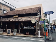 A rustic, traditional building with a sloping tiled roof and wooden exterior sits at a street corner. A signboard with foreign script hangs above the entrance. Several potted plants and a chalkboard menu frame the doorway, suggesting a cafe or small restaurant. Street signs in multiple languages point in different directions, and a few people walk by.