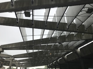 A structure with large metallic beams and panels arranged in a repetitive pattern, creating an industrial and geometric appearance. The image shows a perspective from below, highlighting the intricate design and the play of light and shadow across the surfaces.