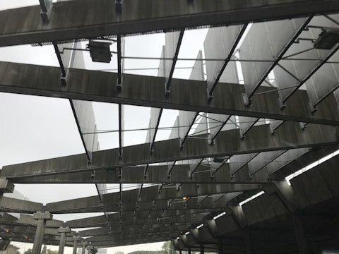 A structure with large metallic beams and panels arranged in a repetitive pattern, creating an industrial and geometric appearance. The image shows a perspective from below, highlighting the intricate design and the play of light and shadow across the surfaces.