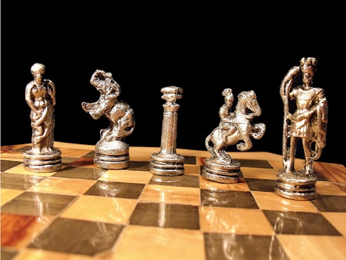 Five ornate, metallic chess pieces resembling classical figures are arranged on a polished wooden chessboard. The pieces include figures of a standing woman, an elephant, a column, a horse with a rider, and a standing man with a staff. The background is a deep black, making the shiny chess pieces stand out prominently.