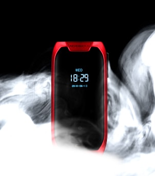 A sleek electronic device with a digital display shows the time '18:29' and the date '2018-06-13.' It is encased in a red frame, with the word 'REVENGER X' visible at the top. Surrounding the device is a swirling cloud of vapor or smoke against a dark background, contrasting with the bright digital screen.