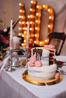 A beautifully decorated layered cake with frosting and pink macarons sits on a gold platter. The cake has a slice removed, revealing a red velvet interior. Nearby, elegant silver candle holders with white and pink candles add a touch of sophistication. In the background, large illuminated letters create a warm, celebratory atmosphere.