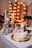 A beautifully decorated layered cake with frosting and pink macarons sits on a gold platter. The cake has a slice removed, revealing a red velvet interior. Nearby, elegant silver candle holders with white and pink candles add a touch of sophistication. In the background, large illuminated letters create a warm, celebratory atmosphere.