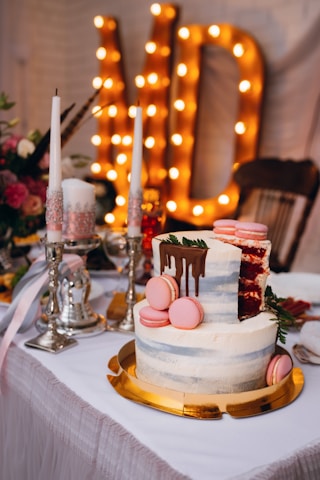 A beautifully decorated layered cake with frosting and pink macarons sits on a gold platter. The cake has a slice removed, revealing a red velvet interior. Nearby, elegant silver candle holders with white and pink candles add a touch of sophistication. In the background, large illuminated letters create a warm, celebratory atmosphere.