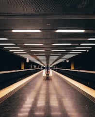 A deserted underground subway station with a symmetrical design, featuring a long, empty platform stretching into the distance. Overhead, fluorescent lights illuminate the tiled floor, creating a sense of depth. The walls and ceiling have a modern metallic texture, and signage is visible but minimal.