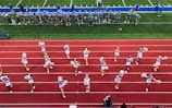 A group of cheerleaders is lined up on a red athletic track, performing routines with pom-poms. Behind them, a football team stands on a green field, dressed in gray and blue jerseys and helmets, observing the scene. Coaches and other officials are stationed along the sidelines.