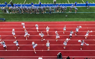 A group of cheerleaders is lined up on a red athletic track, performing routines with pom-poms. Behind them, a football team stands on a green field, dressed in gray and blue jerseys and helmets, observing the scene. Coaches and other officials are stationed along the sidelines.