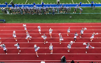 A group of cheerleaders is lined up on a red athletic track, performing routines with pom-poms. Behind them, a football team stands on a green field, dressed in gray and blue jerseys and helmets, observing the scene. Coaches and other officials are stationed along the sidelines.