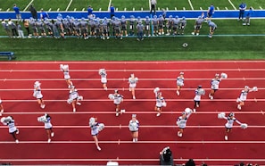 A group of cheerleaders is lined up on a red athletic track, performing routines with pom-poms. Behind them, a football team stands on a green field, dressed in gray and blue jerseys and helmets, observing the scene. Coaches and other officials are stationed along the sidelines.