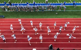 A group of cheerleaders is lined up on a red athletic track, performing routines with pom-poms. Behind them, a football team stands on a green field, dressed in gray and blue jerseys and helmets, observing the scene. Coaches and other officials are stationed along the sidelines.