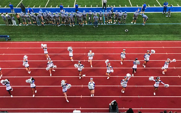 A group of cheerleaders is lined up on a red athletic track, performing routines with pom-poms. Behind them, a football team stands on a green field, dressed in gray and blue jerseys and helmets, observing the scene. Coaches and other officials are stationed along the sidelines.