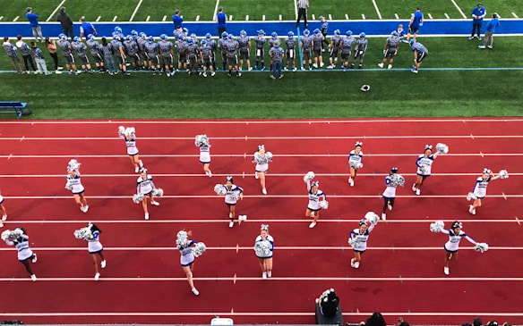 A group of cheerleaders is lined up on a red athletic track, performing routines with pom-poms. Behind them, a football team stands on a green field, dressed in gray and blue jerseys and helmets, observing the scene. Coaches and other officials are stationed along the sidelines.