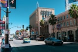 A bustling urban street scene with historic architecture, featuring a wide avenue lined with palm trees and buildings. A theater marquee with vibrant signage is visible. Traffic includes a mix of cars and a bus, and pedestrians walk along the sidewalk. Bright advertisements and billboards are mounted on the buildings, contributing to the lively atmosphere.