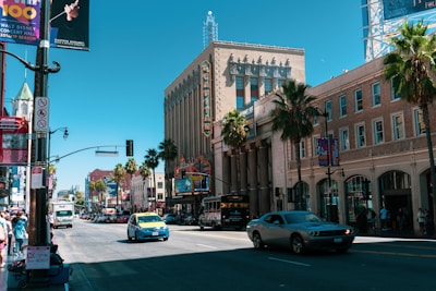 A bustling urban street scene with historic architecture, featuring a wide avenue lined with palm trees and buildings. A theater marquee with vibrant signage is visible. Traffic includes a mix of cars and a bus, and pedestrians walk along the sidewalk. Bright advertisements and billboards are mounted on the buildings, contributing to the lively atmosphere.