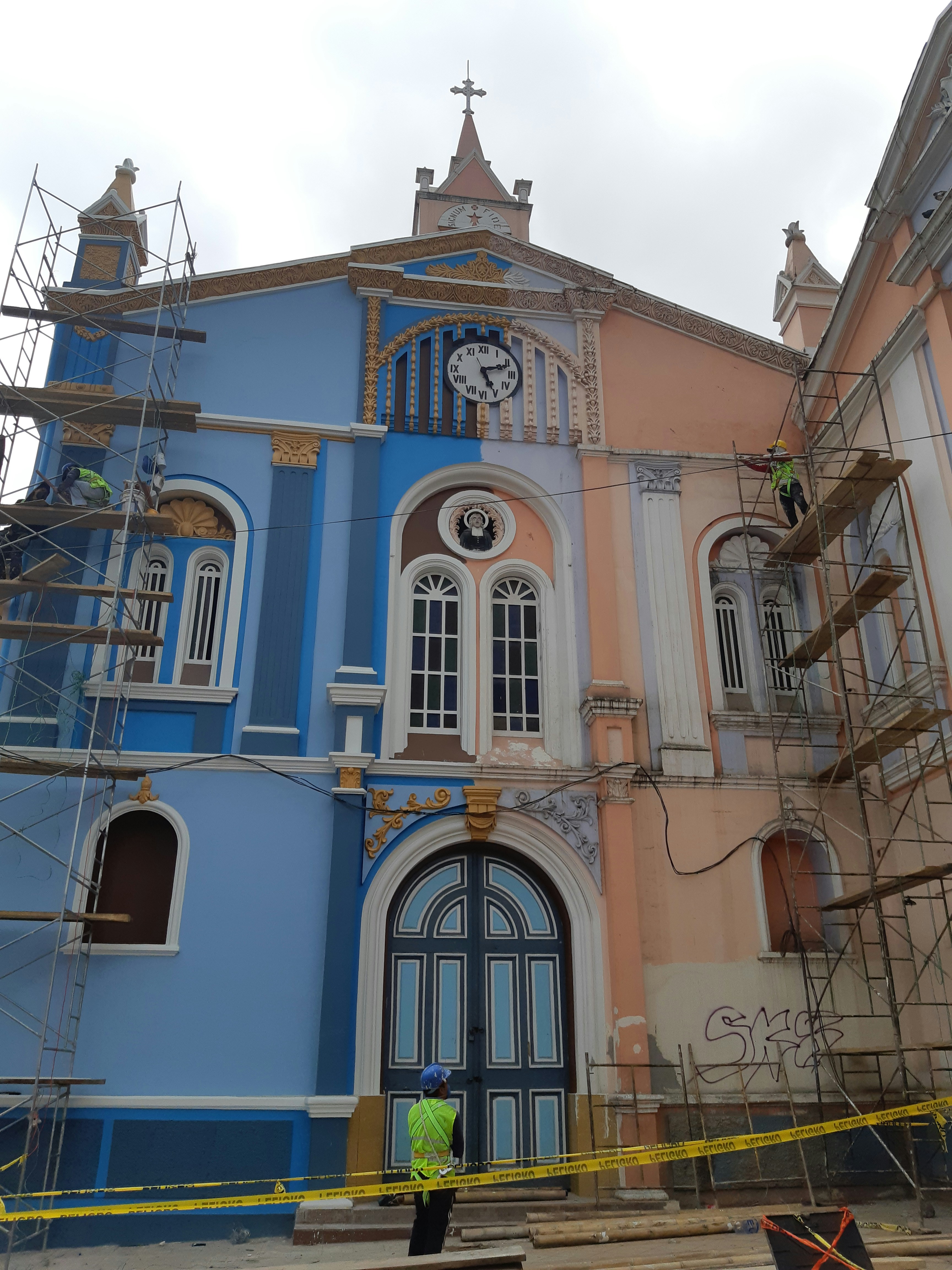 A colorful, historic building with blue and peach hues under renovation. Scaffolding is set up on both sides, and a clock with Roman numerals is visible above the entrance. Ornate details and arched windows enhance the architectural design. A construction worker wearing a helmet and high-visibility vest stands in front of the structure, surrounded by caution tape.