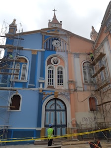 A colorful, historic building with blue and peach hues under renovation. Scaffolding is set up on both sides, and a clock with Roman numerals is visible above the entrance. Ornate details and arched windows enhance the architectural design. A construction worker wearing a helmet and high-visibility vest stands in front of the structure, surrounded by caution tape.