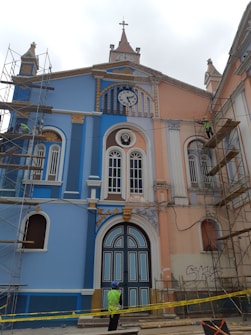 A colorful, historic building with blue and peach hues under renovation. Scaffolding is set up on both sides, and a clock with Roman numerals is visible above the entrance. Ornate details and arched windows enhance the architectural design. A construction worker wearing a helmet and high-visibility vest stands in front of the structure, surrounded by caution tape.