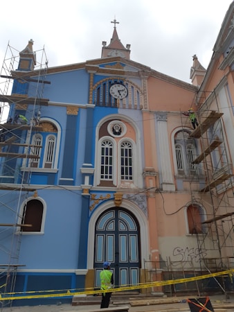 A colorful, historic building with blue and peach hues under renovation. Scaffolding is set up on both sides, and a clock with Roman numerals is visible above the entrance. Ornate details and arched windows enhance the architectural design. A construction worker wearing a helmet and high-visibility vest stands in front of the structure, surrounded by caution tape.