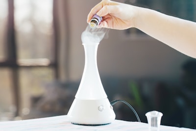 A hand is holding an essential oil bottle over a white, conical diffuser emitting vapor on a table. The background is softly blurred with a large window, creating a serene and calm atmosphere, suggesting a focus on aromatherapy or relaxation.