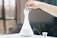 A hand is holding an essential oil bottle over a white, conical diffuser emitting vapor on a table. The background is softly blurred with a large window, creating a serene and calm atmosphere, suggesting a focus on aromatherapy or relaxation.