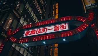 A prominent red and white illuminated sign with Japanese characters is set against the backdrop of a cityscape. The sign features bright lights that are arranged in a glowing border. Nearby, tall buildings with lit windows and additional signage can be seen.