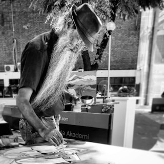 An elderly man with a long beard is engaged in painting outdoors. He wears a hat and glasses and has a paintbrush in one hand and a container in the other. The background includes a tree and a partially visible easel with artwork.