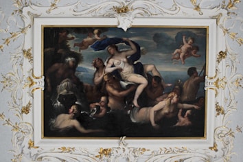 A classical painting depicts a mythological scene with several nude or semi-nude figures. A central figure is draped in fabric, surrounded by others in dynamic poses. Two cherubs are hovering above, amid a cloudy sky. The ornate frame is adorned with intricate gold leaf designs.