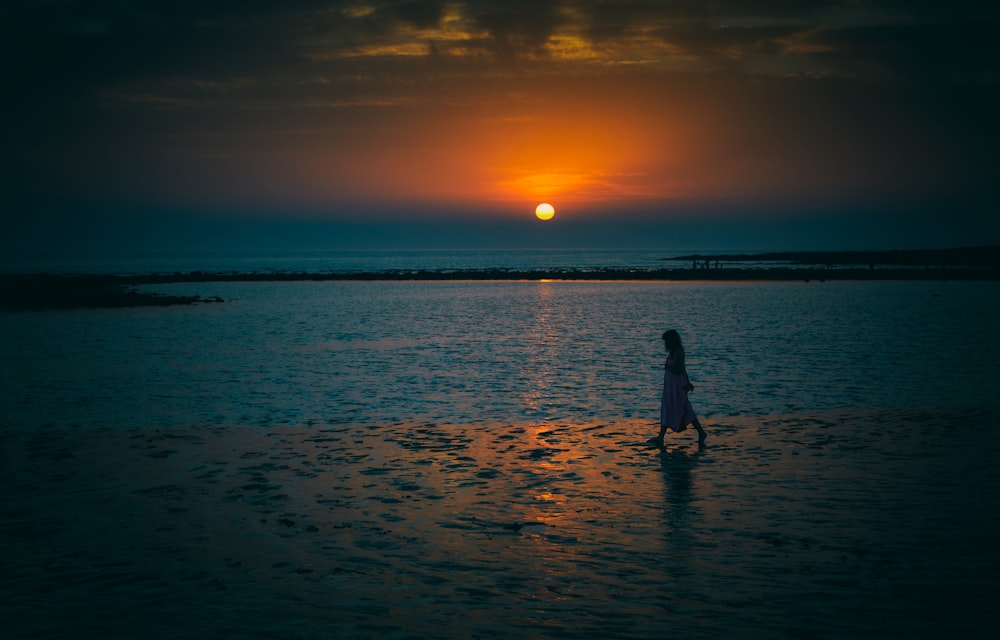 silhouette of woman standing near body of water during sunset