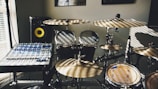 A room with a drum set and a digital audio mixer. Sunlight creates striped patterns across the instruments and control panels. The drum set includes multiple cymbals and drums, and the mixer is equipped with various knobs and sliders. A monitor speaker with a yellow cone is visible in the background.
