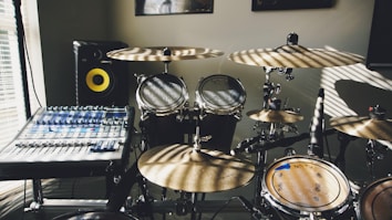 A room with a drum set and a digital audio mixer. Sunlight creates striped patterns across the instruments and control panels. The drum set includes multiple cymbals and drums, and the mixer is equipped with various knobs and sliders. A monitor speaker with a yellow cone is visible in the background.