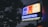 A neon sign with the words 'Downtown Bentonville Inc.' is illuminated against a backdrop of tree leaves and a dim evening sky. The sign features brightly lit building outlines in blue, red, and yellow colors.