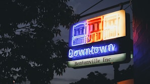 A neon sign with the words 'Downtown Bentonville Inc.' is illuminated against a backdrop of tree leaves and a dim evening sky. The sign features brightly lit building outlines in blue, red, and yellow colors.