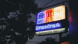 A neon sign with the words 'Downtown Bentonville Inc.' is illuminated against a backdrop of tree leaves and a dim evening sky. The sign features brightly lit building outlines in blue, red, and yellow colors.